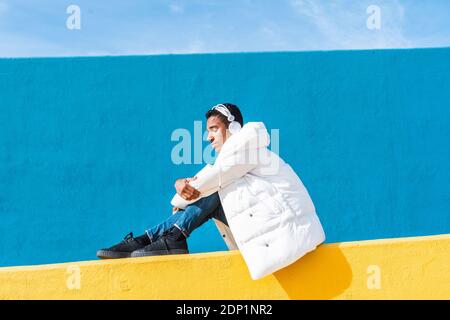 Young trendy man sitting on yellow wall, listening music Stock Photo