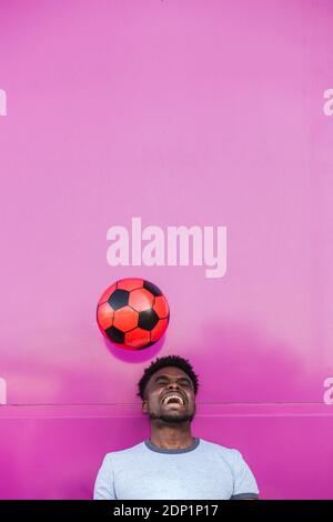 Cheerful young man juggling with soccer ball against pink wall in city Stock Photo