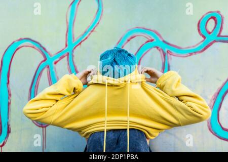 Young woman with dyed blue hair putting on yellow hooded jacket Stock Photo