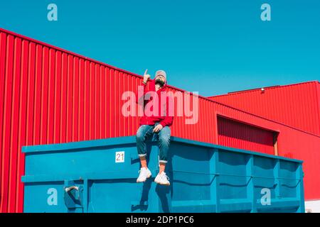 Young man wearing red hoodie, sitting on edge of blue container in front of red wall, raising forefinger Stock Photo