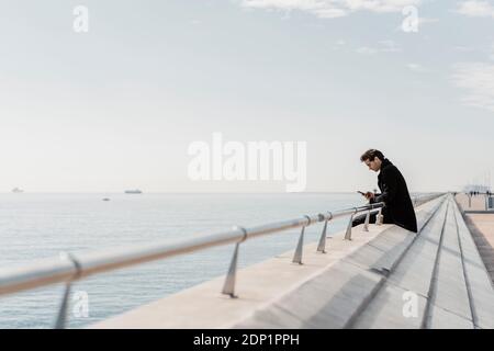Businessman sitting on quay wall checking cell phone Stock Photo