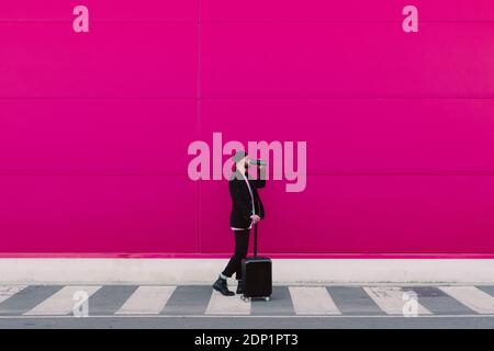 Young man with trolley walking along a pink wall, drinking from a reusable cup Stock Photo