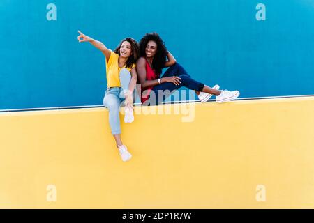 Young woman pointing to smiling friend while sitting on yellow retaining wall Stock Photo