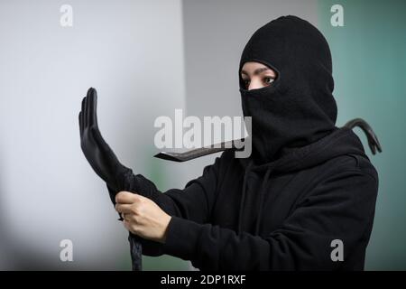 Thief broke into the apartment. House robbery by woman in a black jacket and black mask and crowbar. Burglar in a mask. Thief in a mask trying to break into other people's house Stock Photo