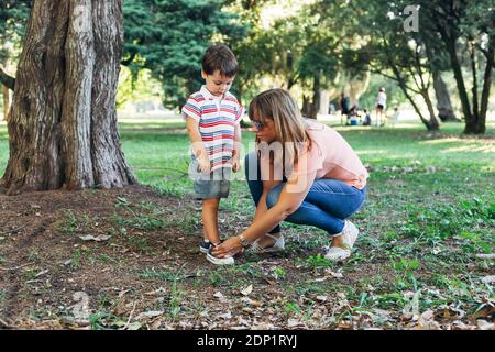 Mom tying sneakers to her little son. Mother with her child in the middle of forest park. Stock Photo