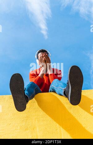 Young man with headphones, sitting on yellow wall, listening music Stock Photo