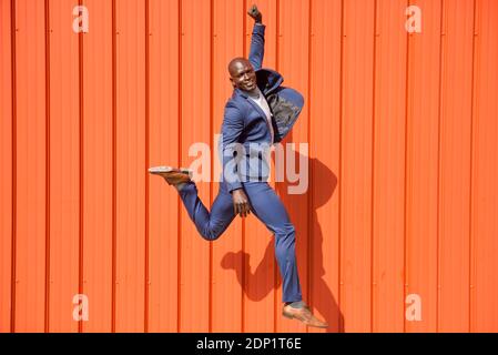 Smiling businessman jumping in the air in front of orange wall Stock Photo