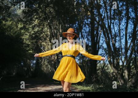 Happy young curly haired woman wearing brown hat and yellow dress walking with open arms and closed eyes Stock Photo