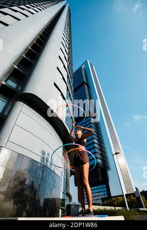 Sporty woman exercising with plastic hoops outside office buildings against blue sky Stock Photo