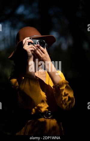 Young woman wearing a brown hat and yellow dress taking a photo with a analog camera in a forest Stock Photo