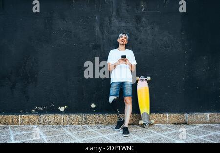 Laughing young man with leg prosthesis and skateboard using smartphone Stock Photo