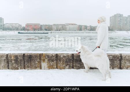 Serbia, Petrovaradin, white dressed young woman standing with white dog in the snow at riverside Stock Photo