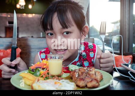 Smile asian child girl eating steak and vegetable salad on the table with holding knife and fork Stock Photo