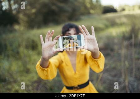 Happy young woman wearing yellow dress taking a white smartphone in front of her face and taking a selfie Stock Photo