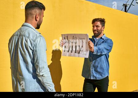 Man holding paper with written text Would You Marry Me on it while standing by yellow wall Stock Photo