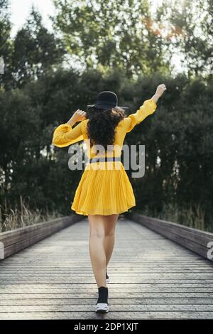 Rear view of a young woman wearing black hat and yellow dress enjoying while is walking along wooden bridge Stock Photo