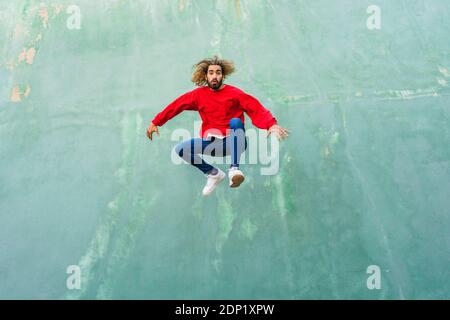 Portrait of young man wearing red sweatshirt jumping in the air in front of green wall Stock Photo