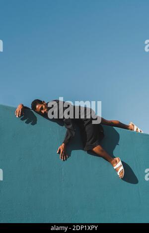 Young man wearing black kaftan lying on blue wall Stock Photo