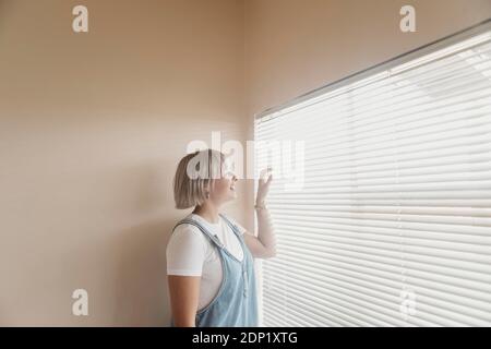Happy young woman looking through blinds at the window Stock Photo