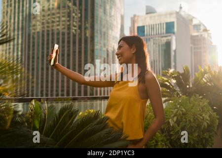 Smiling woman standing on roof terrace in the evening taking selfie with smartphone, Bangkok, Thailand Stock Photo