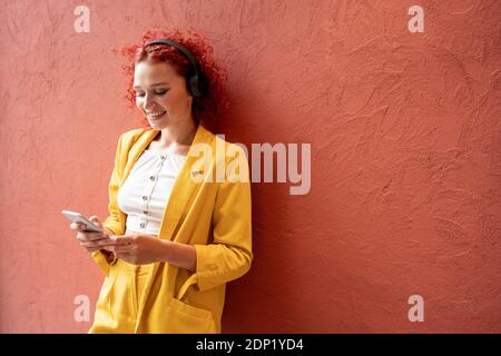 Young woman in yellow suit leaning agaibst red wall, wearing headphones, using smartphone Stock Photo