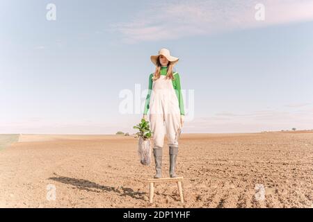 Young woman standing on stool on dry field, carrying plant in a net Stock Photo