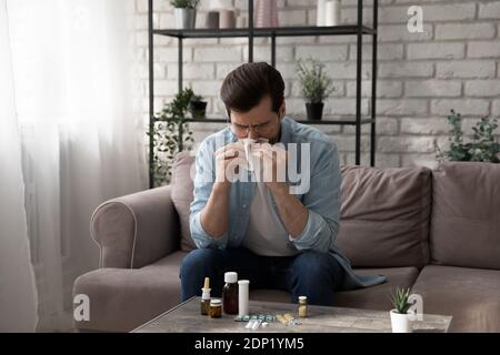 Ill young man sitting on sofa blowing nose taking pills Stock Photo