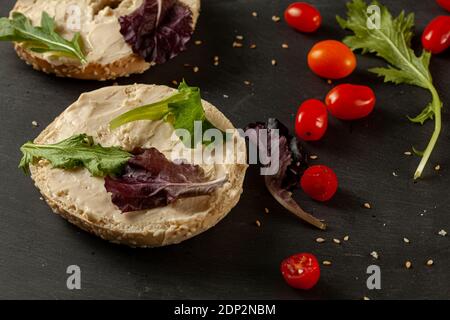 Toasted and sliced sesame bagel with creamy spread on it served with fresh greens (arugula) and cherry tomatoes on black wooden background. A healthy Stock Photo