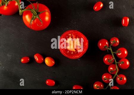 A flat lay image featuring a medley of tomatoes on black wooden background. Some Roma tomatoes and cherry tomatoes are on vine, one of them is sliced Stock Photo