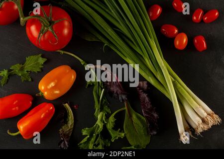 Flat lay image of green leafy vegetable mix, arugula leafs, tomato on vine, bell peppers, cherry tomatoes and a bunch of spring onions on black wooden Stock Photo