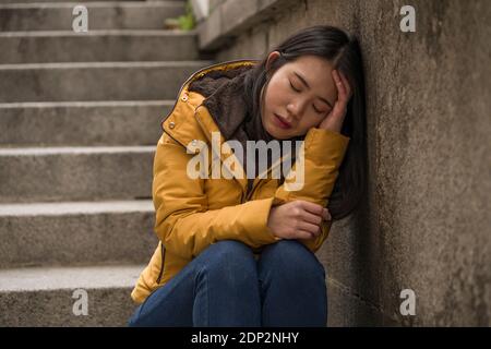 dramatic lifestyle portrait of young attractive sad and depressed Korean woman in winter jacket sitting outdoors on street corner staircase suffering Stock Photo