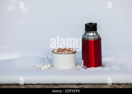 Red thermos next cup with hot chocolate  with marshmallows on table with snow outside