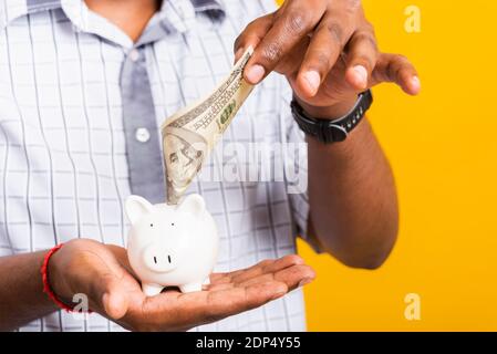 Asian happy portrait young business black man putting dollar bill banknote into a piggy bank, studio shot isolated on white background, Saving deposit Stock Photo