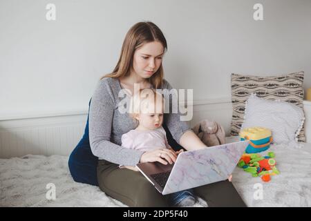 Young Caucasian mother with baby working on laptop from home. Workplace of freelance woman student with kid toddler. Stay at home single mom earning Stock Photo