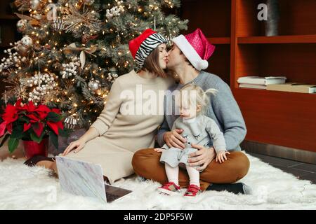 Kissing mother and father in Santa hats with baby girl calling family friends on video call. Distant remote holiday celebration online on Internet. Fa Stock Photo