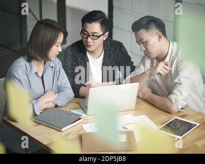 team of three young asian entrepreneurs working together in office discussing business plan using laptop Stock Photo