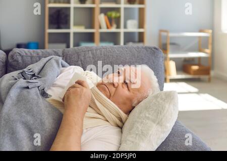 Sad senior man in scarf lying in bed with handkerchief under blanket and feeling ill Stock Photo