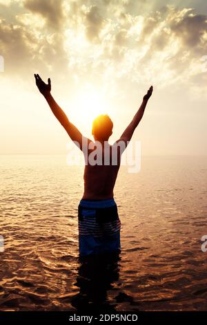 Toned Photo of Man Silhouette with Hands Up in the Water on the Sunset Background Stock Photo