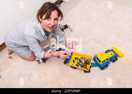 Exhausted and elegant dressed business woman is sitting on the floor and cleaning the toys of her kids. She looks unhappy and frustrated Stock Photo