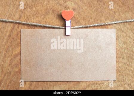 Blank postcard on a rope with a clothespin in the shape of a heart on a wooden background, Valentine's Day theme. Stock Photo