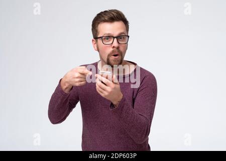 Caucasian man using smartphone pointing at screen, texting message. Millennial guy social network user hold cell phone. Stock Photo