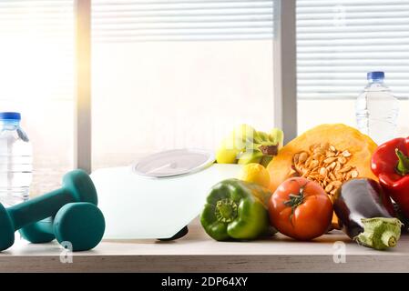 Fruits and vegetables on a kitchen table along with dumbbells and scales for healthy living and diet with window background. Front view. Horizontal co Stock Photo