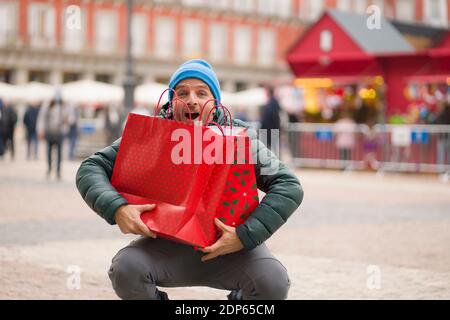 Christmas shopping in town - urban lifestyle portrait of happy and excited man in winter hat carrying shopping bags at xmas market smiling cheerful an