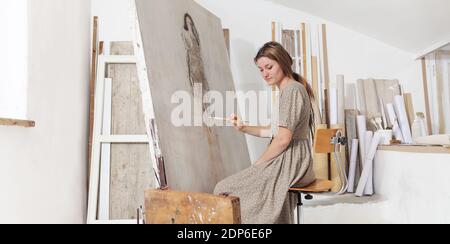 Indoor shot of young female artist sitting in front of the canvas and painting in bright white studio wearing bohemian chic clothin Stock Photo