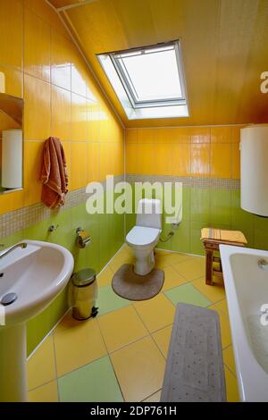 Interior of modern bathroom and toilet in attic room. Bath, toilet, trash can, hot water boiler mirror, dormer window, yellow and green ceramic tiles Stock Photo