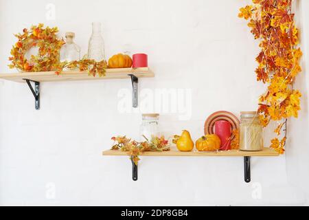Different multicolored decorative pumpkins with yellow leaves on a wooden shelf of white wall. Autumn or fall home interior decor. Stock Photo