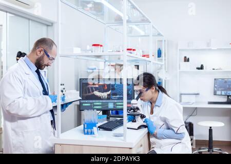 Chemist wearing safety glasses and using tablet pc in lab. Team of researchers doing pharmacology engineering in sterile laboratory for healthcare industry with african assistant in the background. Stock Photo