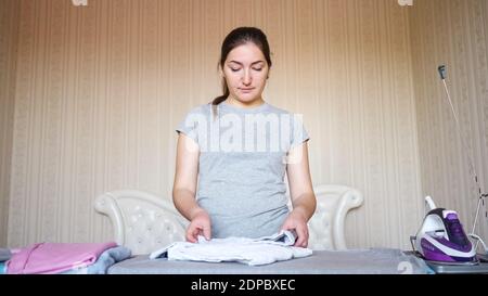 Relaxed housewife with dark hair stacks clothes after ironing with modern purple electric iron on special board close view Stock Photo