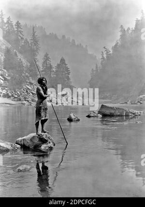 Iconic vintage photograph by Edward Curtis entitled A Smoky Day at the Sugar Bowl A Hupa man stands on a rock in a lake with a forest background. Stock Photo