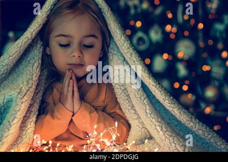 Kid Making Magic Wish for Winter Holidays. Baby Near the Christmas Tree at Home. Under the Plaid Praying. Waiting for Santa at Night. Stock Photo